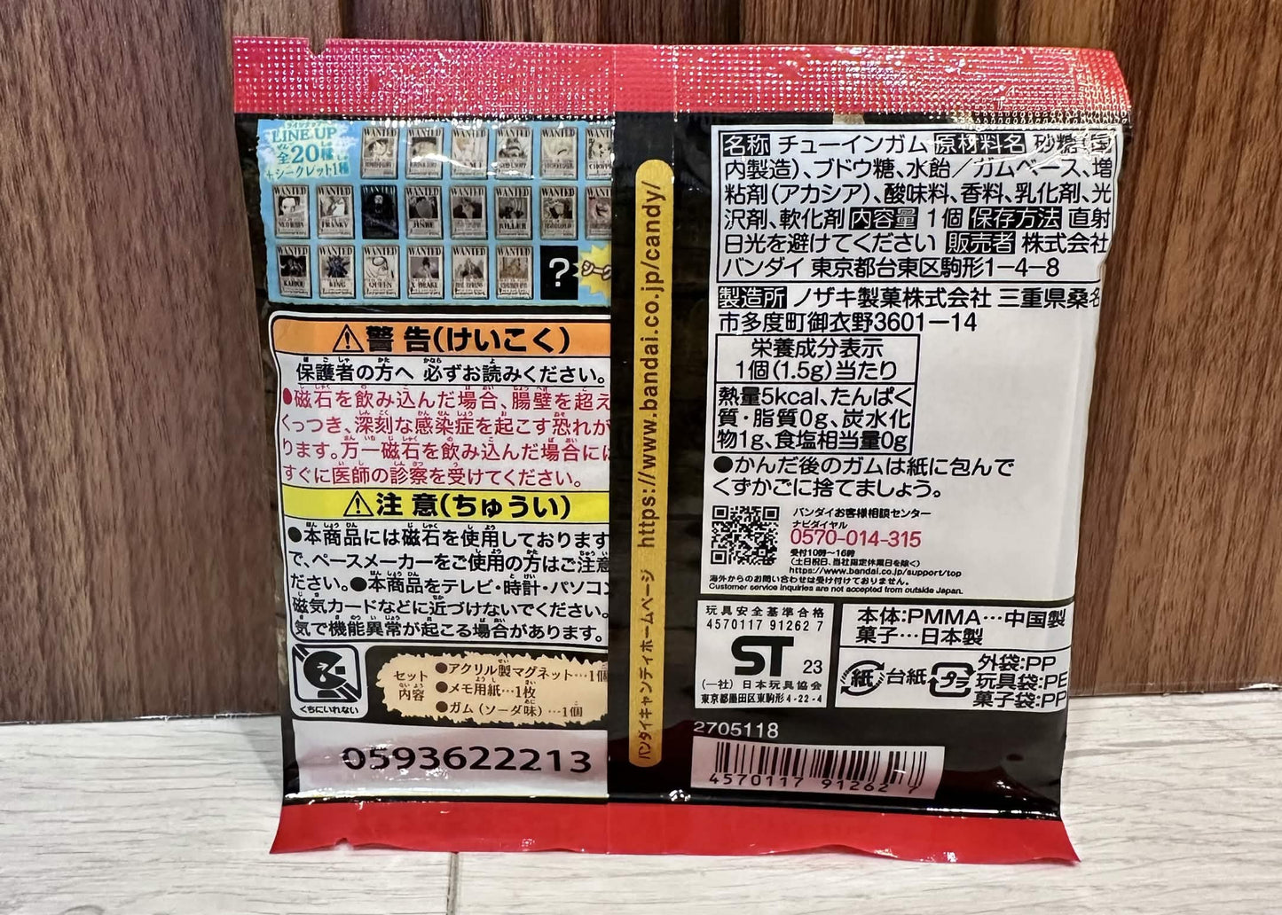 The back of a Japanese toy packaging blister pack for 'One Piece Chara-Mangets' by Bandai Namco, resting on a light wood floor against a dark wood panel background.